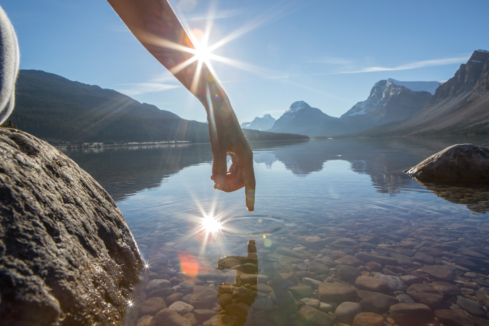 Finger touches surface of mountain lake. The sun and the landscape is reflecting on the water.